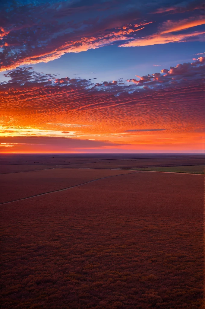 A breathtaking sunset over the expansive plains of eastern New Mexico, Llano Estacado. The sky is painted with vibrant hues of orange, pink, and purple, casting a warm glow over the vast, open landscape. Wispy clouds add texture to the sky, while the flat terrain stretches endlessly, dotted with sparse vegetation typical of the region. The scene captures the serene and majestic beauty of the high plains at dusk.