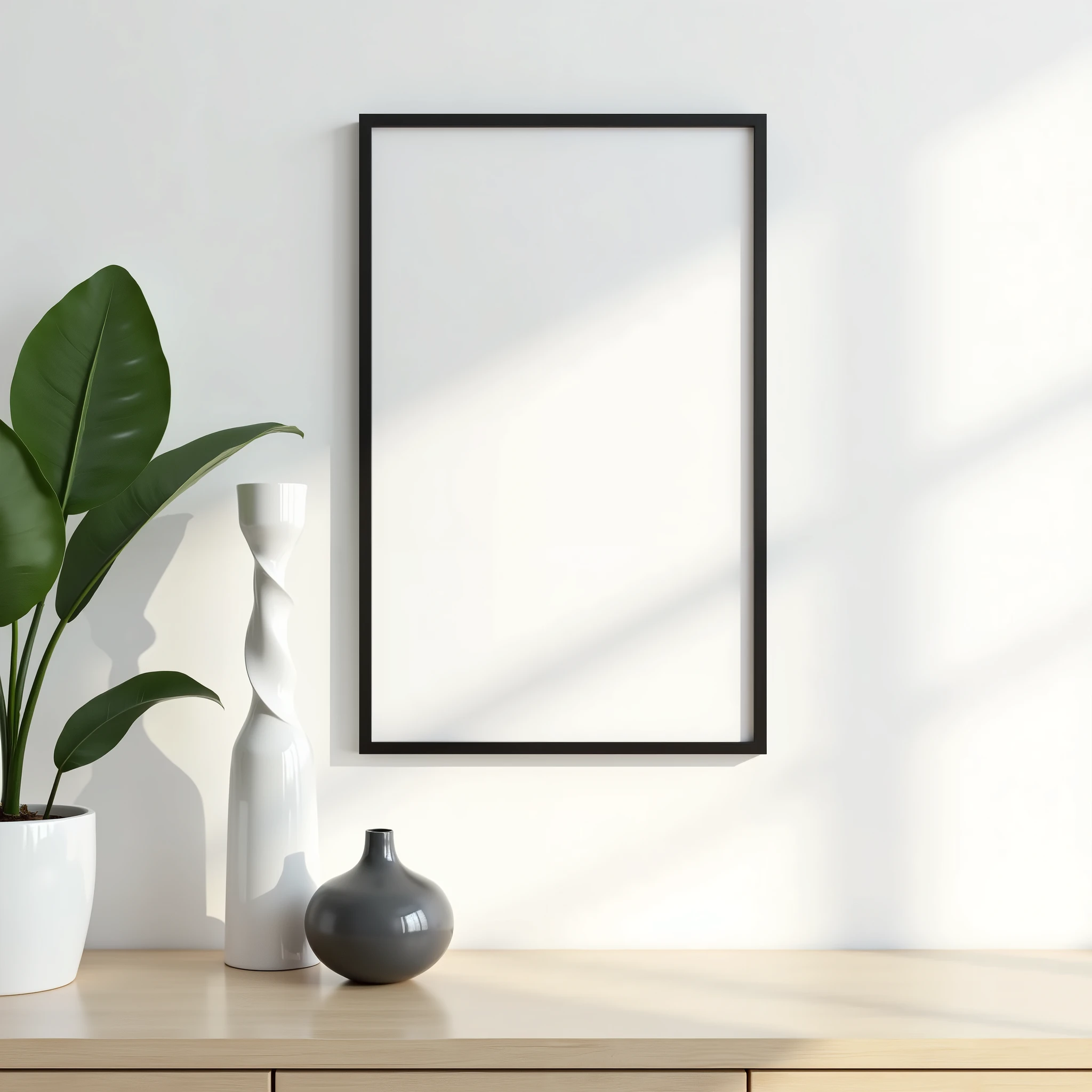 A modern minimalist interior scene featuring a white wall with a single black-framed A4-sized picture frame hung slightly off-center. Below the frame is a light wooden surface, which could be a tabletop or a shelf. On this surface, there are two vases: one tall and sleek in a glossy white, with a subtle twisted design, and another smaller, more rounded vase in a glossy dark gray. To the left of the scene, a large green houseplant with broad leaves partially enters the frame, adding a touch of nature. The scene is softly lit by natural light, creating a clean and calming atmosphere with gentle shadows