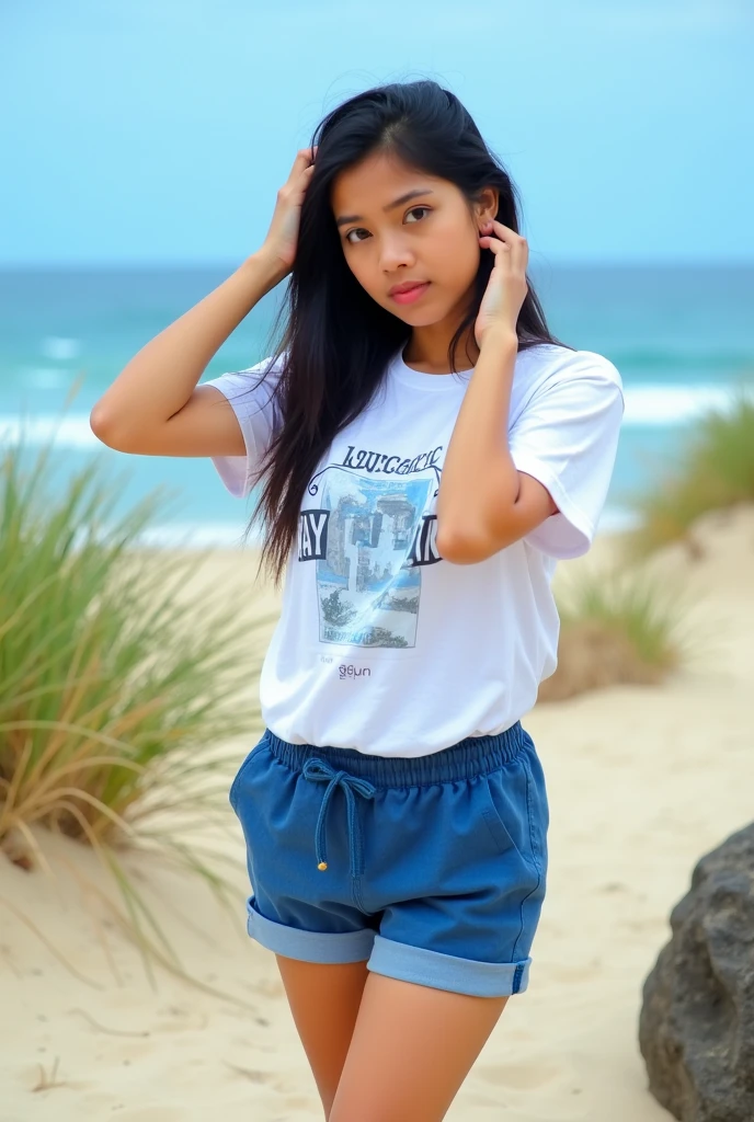 The image shows a young woman standing on a sandy beach with the ocean in the background. She is wearing a white t-shirt with a graphic design on it and blue shorts with a drawstring waistband. She has long dark hair and is posing with one hand on her hip and the other resting on her head. The woman is looking directly at the camera with a serious expression on her face. The sky is blue and the ocean is visible in the distance, with the waves crashing onto the shore. The beach is surrounded by tall grasses and there is a large rock in the foreground. The overall mood of the image is peaceful and serene.