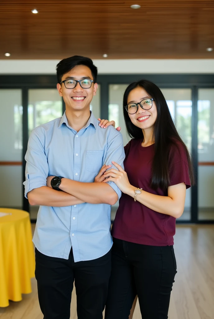 Image is a vertical, full-body photograph of two Filipino people standing indoors. The person on the left is a young man with short black hair, wearing glasses, a light blue shirt, black pants, and a watch on his left wrist. He has a light skin tone and is smiling with his arms crossed. The person on the right is a young woman with long black hair, wearing glasses, a maroon t-shirt, and black pants. She has a light skin tone and is smiling while touching the man's arm. The background features a modern interior with a wooden ceiling, glass doors, and a yellow tablecloth on a table to the left.