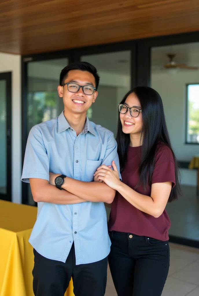 Image is a vertical, full-body photograph of two Filipino people standing indoors. The person on the left is a young man with short black hair, wearing glasses, a light blue shirt, black pants, and a watch on his left wrist. He has a light skin tone and is smiling with his arms crossed. The person on the right is a young woman with long black hair, wearing glasses, a maroon t-shirt, and black pants. She has a light skin tone and is smiling while touching the man's arm. The background features a modern interior with a wooden ceiling, glass doors, and a yellow tablecloth on a table to the left.