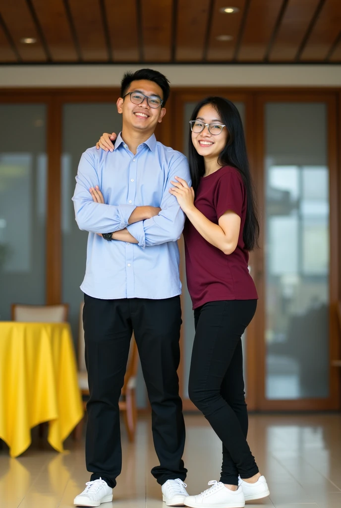 Image is a vertical, full-body photograph of two Filipino people standing indoors. The person on the left is a young man with short black hair, wearing glasses, a light blue shirt, black pants, a watch on his left wrist, and white shoes. He has a light skin tone and is smiling with his arms crossed. The person on the right is a young woman with long black hair, wearing glasses, a maroon t-shirt, black pants, and white shoes. She has a light skin tone and is smiling while touching the man's arm. The background features a modern interior with a wooden ceiling, glass doors, and a yellow tablecloth on a table to the left.
