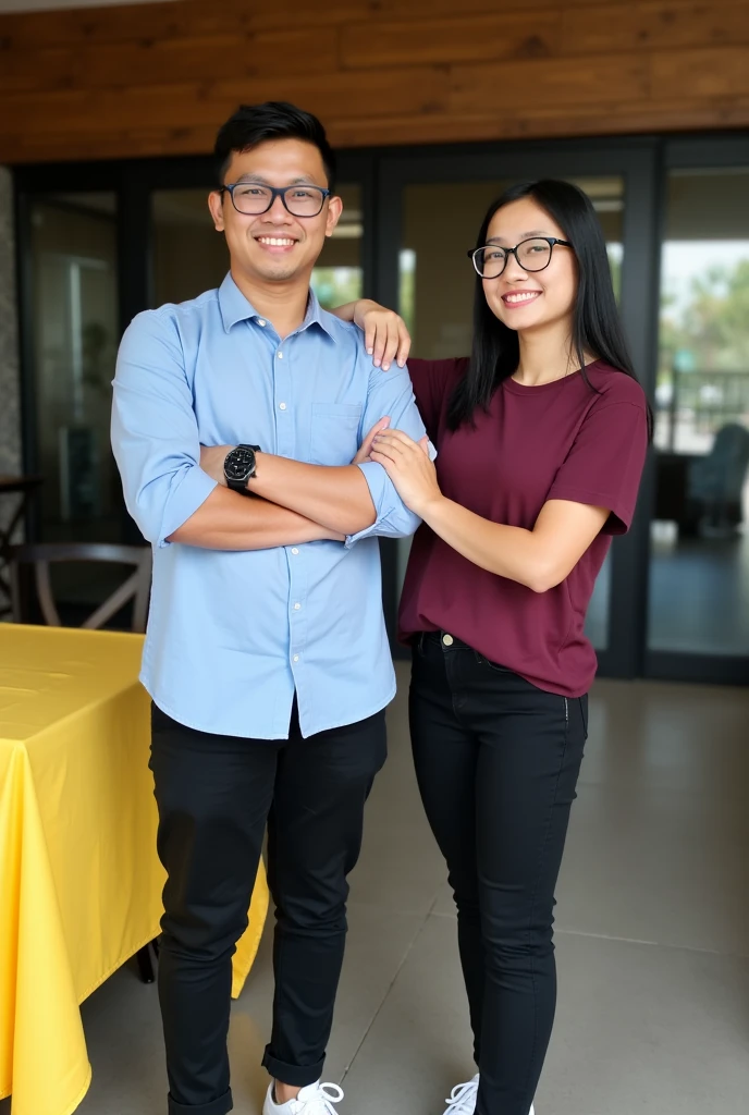 Image is a vertical, full-body photograph of two Filipino people standing indoors. The person on the left is a young man with short black hair, wearing glasses, a light blue shirt, black pants, a watch on his left wrist, and white shoes. He has a light skin tone and is smiling with his arms crossed. The person on the right is a young woman with long black hair, wearing glasses, a maroon t-shirt, black pants, and white shoes. She has a light skin tone and is smiling while touching the man's arm. The background features a modern interior with a wooden ceiling, glass doors, and a yellow tablecloth on a table to the left.
