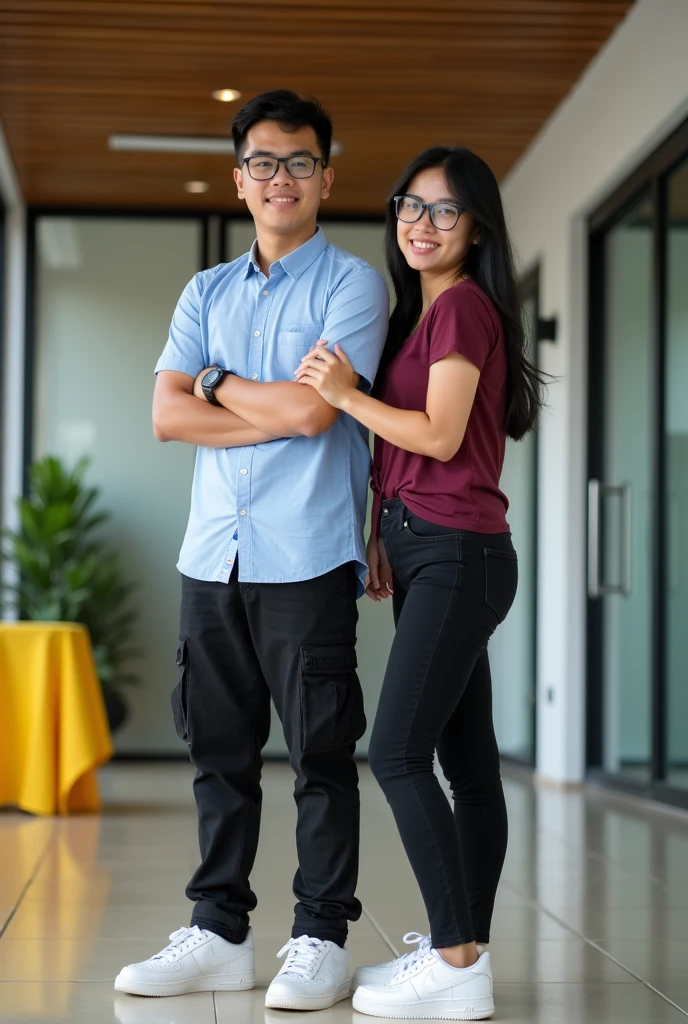Image is a vertical, full-body photograph of two Filipino people standing indoors. The person on the left is a young man with short black hair, wearing glasses, a light blue shirt, black cargo pants, a watch on his left wrist, and white samba shoes. He has a light skin tone and is smiling no teeth with his arms crossed. The person on the right is a young woman with long black hair, wearing glasses, a maroon t-shirt, black skinny jeans, and white nike airforce shoes. She has a lighter skin tone and is slightly smiling while touching the man's arm. The background features a modern interior with a wooden ceiling, glass doors, and a yellow tablecloth on a table to the left.