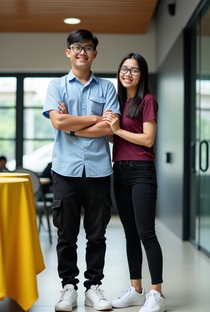 Image is a vertical, full-body photograph of two Filipino people standing indoors. The person on the left is a young man with short black hair, wearing glasses, a light blue shirt, black cargo pants, a watch on his left wrist, and white samba shoes. He has a light skin tone and is smiling no teeth with his arms crossed. The person on the right is a young woman with long black hair, wearing glasses, a maroon t-shirt, black skinny jeans, and white nike airforce shoes. She has a lighter skin tone and is slightly smiling while touching the man's arm. The background features a modern interior with a wooden ceiling, glass doors, and a yellow tablecloth on a table to the left.