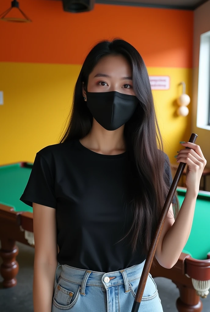 Image is a candid photograph of a young woman standing in an indoor recreational area, likely a pool hall. She has long, straight black hair and is wearing a black face mask that covers her nose and mouth. Her skin tone is light to medium. She is dressed in a black t-shirt, light blue high-waisted jeans, and white shoes. She is holding a pool cue in her right hand and appears to be smiling, as indicated by her eyes. The background features a pool table with green felt, and the walls are painted in bright orange and yellow colors. There is a 'No Smoking' sign visible on the wall behind her.