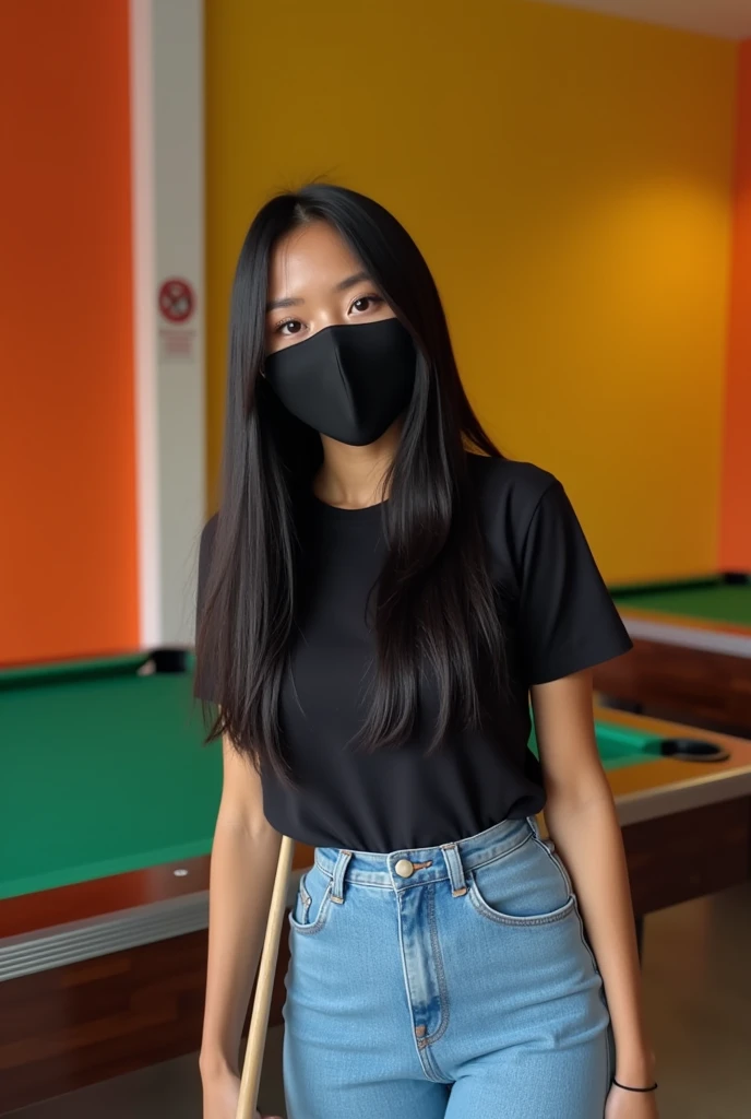 Image is a candid photograph of a young woman standing in an indoor recreational area, likely a pool hall. She has long, straight black hair and is wearing a black face mask that covers her nose and mouth. Her skin tone is light to medium. She is dressed in a black t-shirt, light blue high-waisted jeans, and white shoes. She is holding a pool cue in her right hand and appears to be smiling, as indicated by her eyes. The background features a pool table with green felt, and the walls are painted in bright orange and yellow colors. There is a 'No Smoking' sign visible on the wall behind her.
