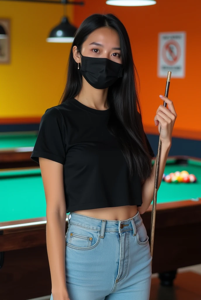 Image is a candid photograph of a young woman standing in an indoor recreational area, likely a pool hall. She has long, straight black hair and is wearing a black face mask that covers her nose and mouth. Her skin tone is light to medium. She is dressed in a black t-shirt, light blue high-waisted jeans, and white shoes. She is holding a pool cue in her right hand and appears to be smiling, as indicated by her eyes. The background features a pool table with green felt, and the walls are painted in bright orange and yellow colors. There is a 'No Smoking' sign visible on the wall behind her.