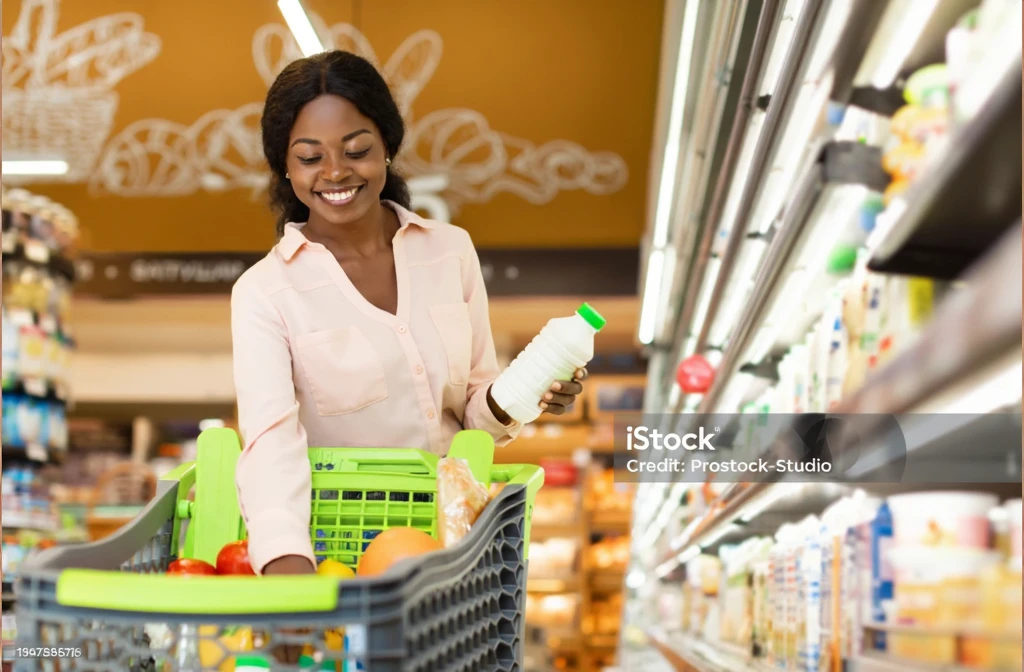 The photo shows a brunette woman standing in the aisle of a supermarket, holding a bag of beans. She is in front of a gondola with various items on display. In the foreground is a shopping cart containing a few items, including what appears to be bread and possibly other groceries.