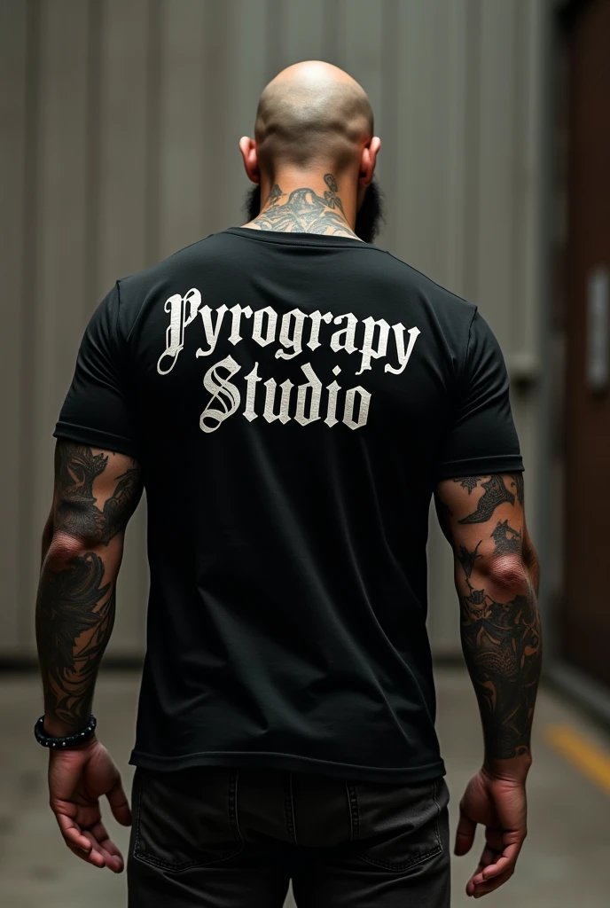 Shaved, dark, tattooed man from behind wearing a black t-shirt with the legend Pyrograpy Studio 

