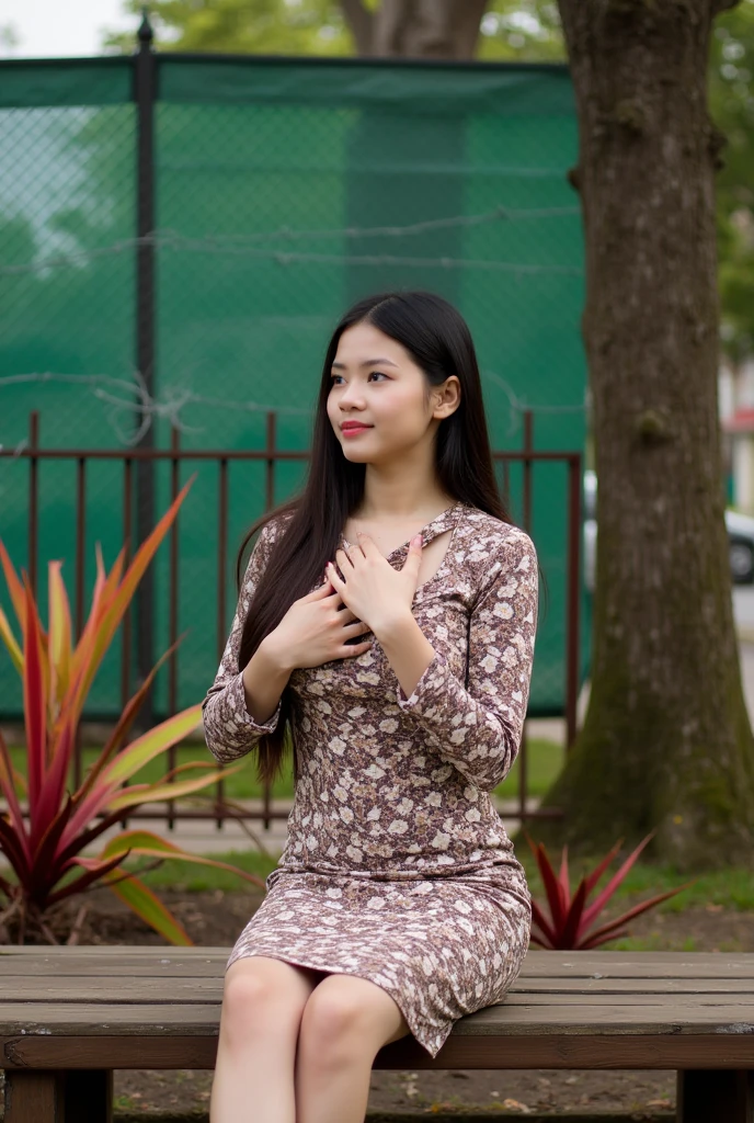 portrait-style photograph featuring a young woman seated on a wooden bench. The layout is vertical, with the subject positioned slightly to the right of the center. She has long, dark hair cascading over her shoulders and is wearing a fitted, long-sleeved dress with a brown and white floral pattern. Her skin is light, and she has a calm expression with her right hand placed gently on her chest. In the background, there is a tree trunk to her right, and a fence with barbed wire and green tarpaulin behind it. There are also some plants with red and green leaves near the fence, adding a touch of nature to the scene.
