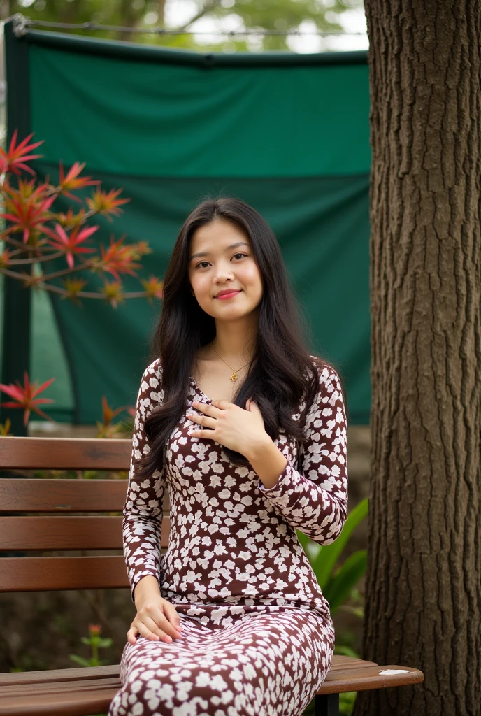 portrait-style photograph featuring a young woman seated on a wooden bench. The layout is vertical, with the subject positioned slightly to the right of the center. She has long, dark hair cascading over her shoulders and is wearing a fitted, long-sleeved dress with a brown and white floral pattern. Her skin is light, and she has a calm expression with her right hand placed gently on her chest. In the background, there is a tree trunk to her right, and a fence with barbed wire and green tarpaulin behind it. There are also some plants with red and green leaves near the fence, adding a touch of nature to the scene.