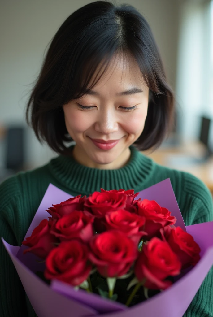 A close-up portrait of a woman with short dark hair, holding a bouquet of red roses wrapped in purple paper. The woman is wearing a green sweater and is looking down at the flowers with a gentle smile. The background is blurred, showing a room with light walls and some office furniture in the distance.japanese,40years old