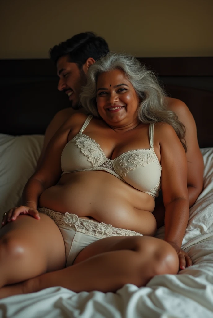 A beautiful gorgeous plump Indian 60 woman wearing a bra panty, long hair, huge breasts, hot body curves, smiley face, white skin, laying on bed with 25 years handsome man