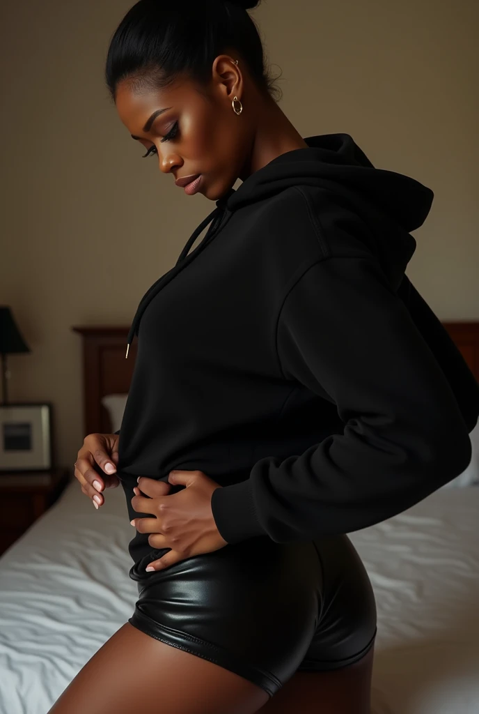 a black woman with her hair in a bun, wearing a black hoodie, bending over and pulling up her black leather shorts, in her bedroom getting dressed