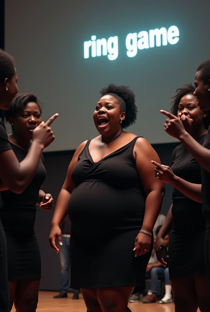 A black woman in a play on stage chubby with her l hair in a low bun in a black dress acting shocked while 5 black people in black clothes on stage point at her with their mouth covered while the audience is around them with the words RING GAME on the screen
