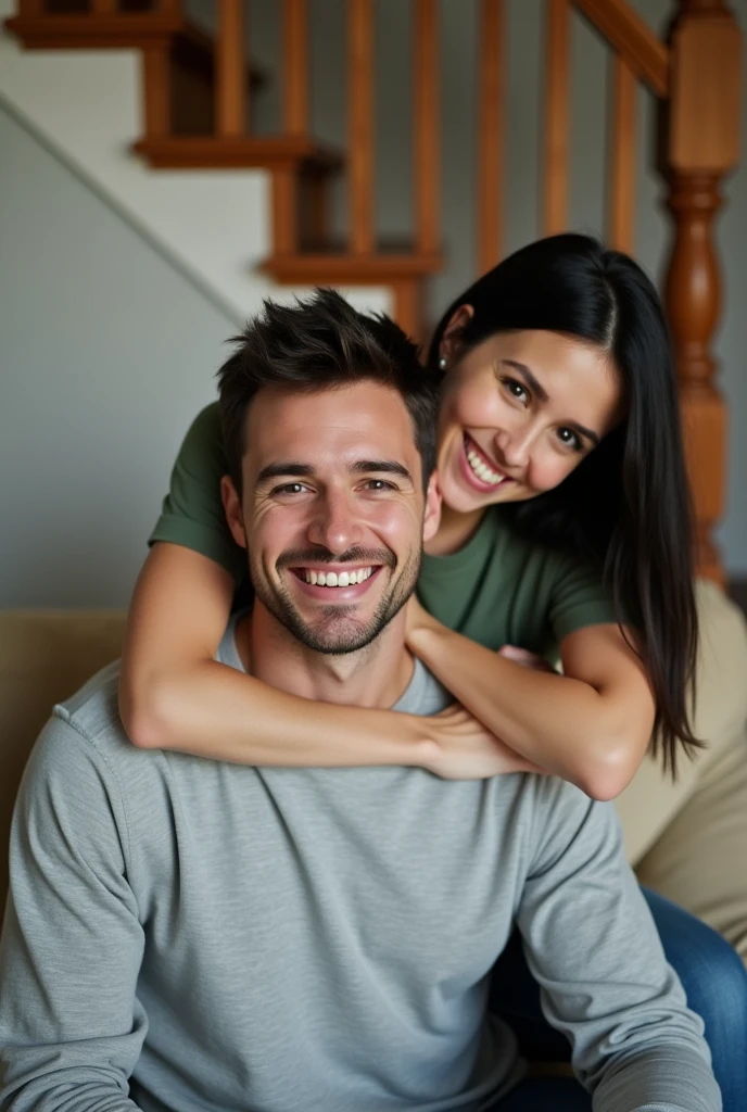 Candid, indoor photograph featuring a young couple in a cozy home setting. The man, with short dark hair is seated on a beige couch, wearing a light gray long-sleeve shirt. The woman, with long straight black hair, is leaning over his shoulders, embracing him from behind, and smiling warmly. She is dressed in a casual green t-shirt and jeans. The background includes a staircase with wooden railings and a partially visible room, adding to the homely atmosphere. The lighting is soft, creating a warm and inviting ambiance.