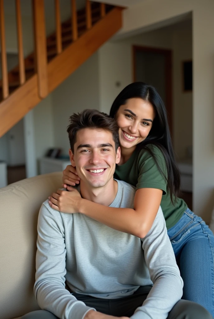 Candid, indoor photograph featuring a young couple in a cozy home setting. The man, with short dark hair is seated on a beige couch, wearing a light gray long-sleeve shirt. The woman, with long straight black hair, is leaning over his shoulders, embracing him from behind, and smiling warmly. She is dressed in a casual green t-shirt and jeans. The background includes a staircase with wooden railings and a partially visible room, adding to the homely atmosphere. The lighting is soft, creating a warm and inviting ambiance.