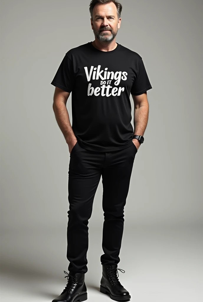 Ricky Gervais wearing a black shirt and black pants and black boots and wearing a shirt with that reads Vikings Do It Better 