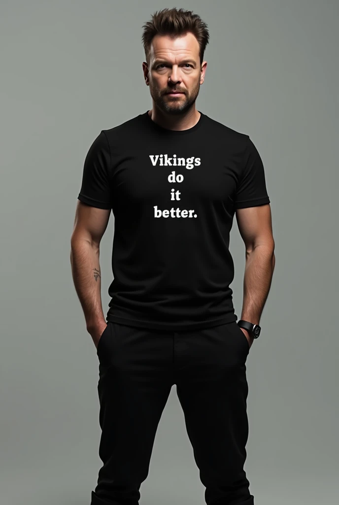 Ricky Gervais wearing a black shirt and black pants and black boots and wearing a shirt with that reads Vikings Do It Better Close up no beard and mustache