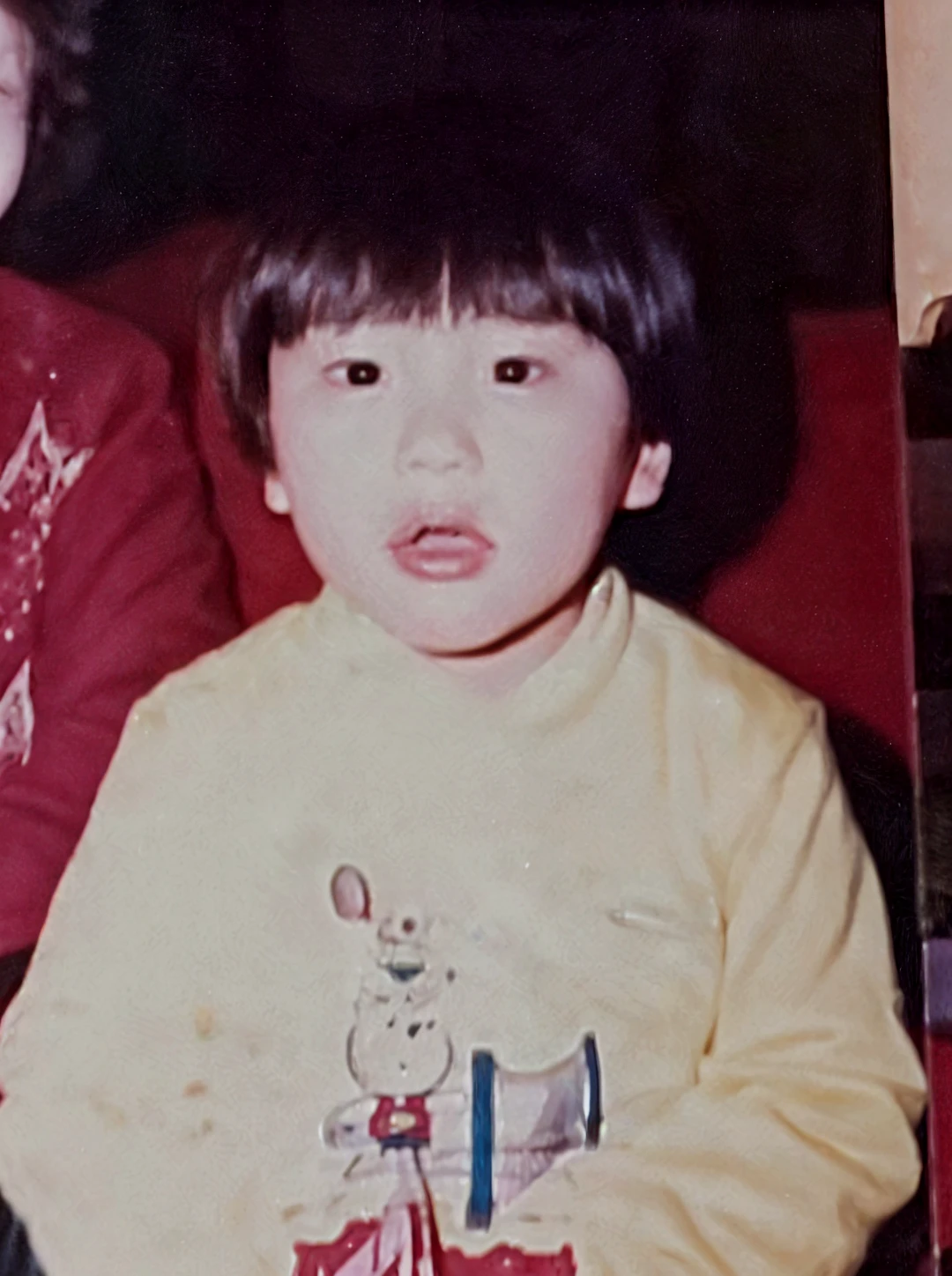 The image is a vintage photo of a young boy in a white long sleeve sweater, standing in front of a white cabinet, with a dark-colored floor and a glass door in the background.