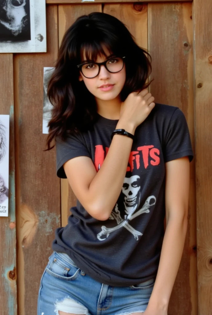 This is a color photograph of a young woman named phoebe standing against a wooden background with band posters. She has shoulder-length, wavy, dark brown hair with bangs that frame her face. Her facial expression is neutral, but intensely looking at the viewer, one eyebrow raised. She is wearing thick rimmed glasses. She is wearing a dark gray, short-sleeved t-shirt made of a soft, slightly textured fabric. She has a medium bust. The t-shirt features a graphic on the front. The graphic is the band Misfits skull logo and name. The woman is also wearing ripped blue, medium-wash denim shorts that fit her comfortably, and shows a bit of her legs. Her right hand is gently touching her neck, and her wrist has black wristband and her left hand is resting by her side. The overall style of the photograph is casual, emphasizing the fashion element with a focus on the t-shirt graphic. The image is well-lit, with no harsh shadows, ensuring that all details are clearly visible.