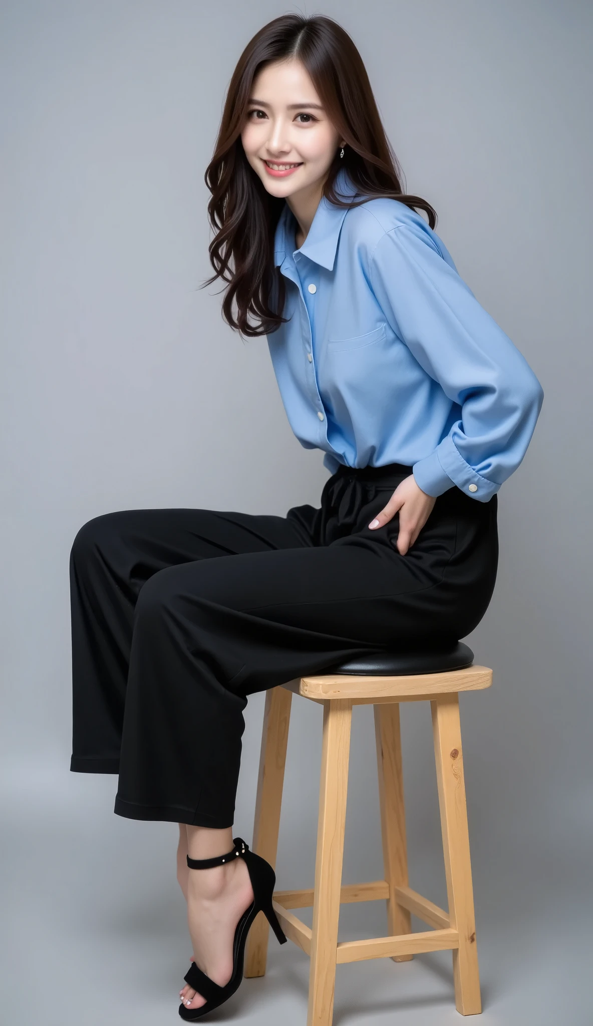 The image is a portrait of a asian young woman sitting on a wooden stool. She is wearing a blue collared shirt and black pants that are loose-fitting and have a high waist. The person's legs are bent at the knees and their left leg is lifted up, as if they are about to sit on the stool. They are wearing black high-heeled sandals. The stool is made of wood and has a black cushion on the backrest. The background is a plain grey color. The woman has long dark hair that is falling over her shoulders and is smiling at the camera. She appears to be in a relaxed pose, with one leg slightly bent at an angle and the other extended up in the air.