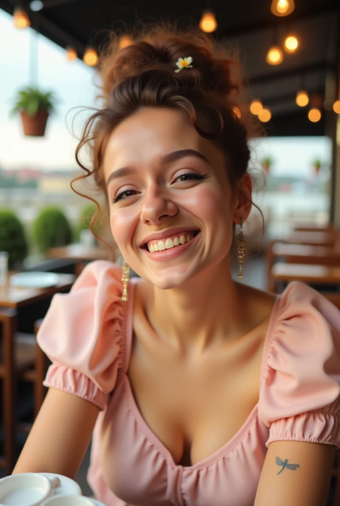 The image is a photograph capturing a candid moment of a woman seated at an outdoor table, likely at a restaurant or café. The woman, with light skin and curly, light brown hair styled in a messy bun, is wearing a soft pink, short-sleeved blouse with ruffled shoulders. She has a tattoo of a dragonfly on her right arm. Her hair is adorned with small, delicate, floral hairpins.