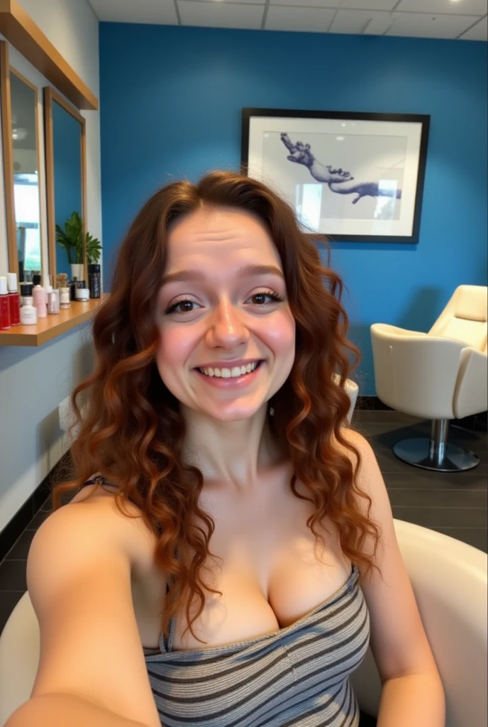 The image is a photograph taken in a modern, well-lit salon. A young woman with long, curly auburn hair is seated in a salon chair, taking a selfie with her smartphone. She has fair skin and wears a sleeveless top with a horizontal stripe pattern. The salon has a contemporary design with a blue wall in the background adorned with framed artwork and a shelf holding various hair care products.