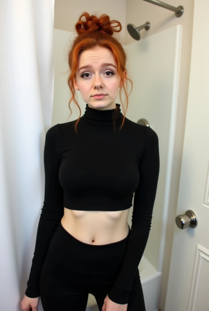 The image is a photograph of a young woman taken in a bathroom. She is standing in front of a white shower curtain and a white door with a silver doorknob. The woman has light skin and long, curly red hair tied up in a messy bun on top of her head. She is wearing a black long-sleeve crop top that reveals a small portion of her midriff, and black high-waisted leggings.