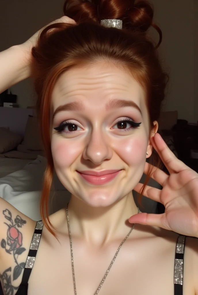 The image is a high-resolution photograph of a young woman taking a selfie in what appears to be a cozy, indoor setting. She is a Caucasian woman with fair skin, freckles, and auburn hair styled in a loose updo with a metallic hair tie. Her facial expression is playful, with a slight smile and raised eyebrows, and she is wearing dark, bold makeup including eyeliner and lipstick, shinestrap showing.