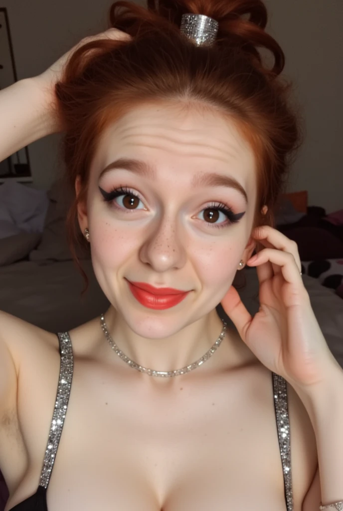 The image is a high-resolution photograph of a young woman taking a selfie in what appears to be a cozy, indoor setting. She is a Caucasian woman with fair skin, freckles, and auburn hair styled in a loose updo with a metallic hair tie. Her facial expression is playful, with a slight smile and raised eyebrows, and she is wearing dark, bold makeup including eyeliner and lipstick, shinestrap showing.