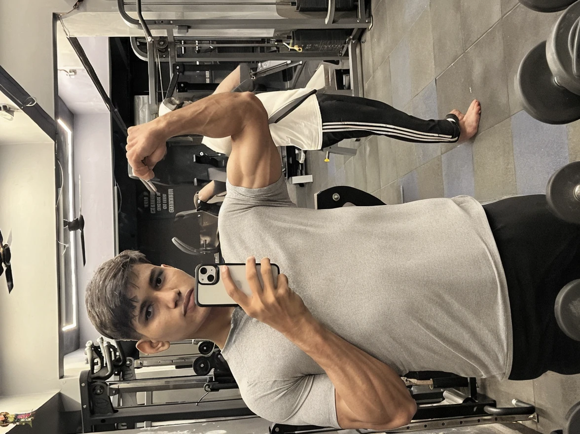 a boy using a bodybuilding machine in a gym, capturing a real image with his phone (Samsung S22 Ultra) in the mirror. The boy has six packs and weighs 65 kg. There is another person present in the image, and the boy has a toned physique without being overly muscular. (best quality, highres), (realistic:1.37), detailed muscles, athletic posture, gym equipment, natural lighting, vibrant colors