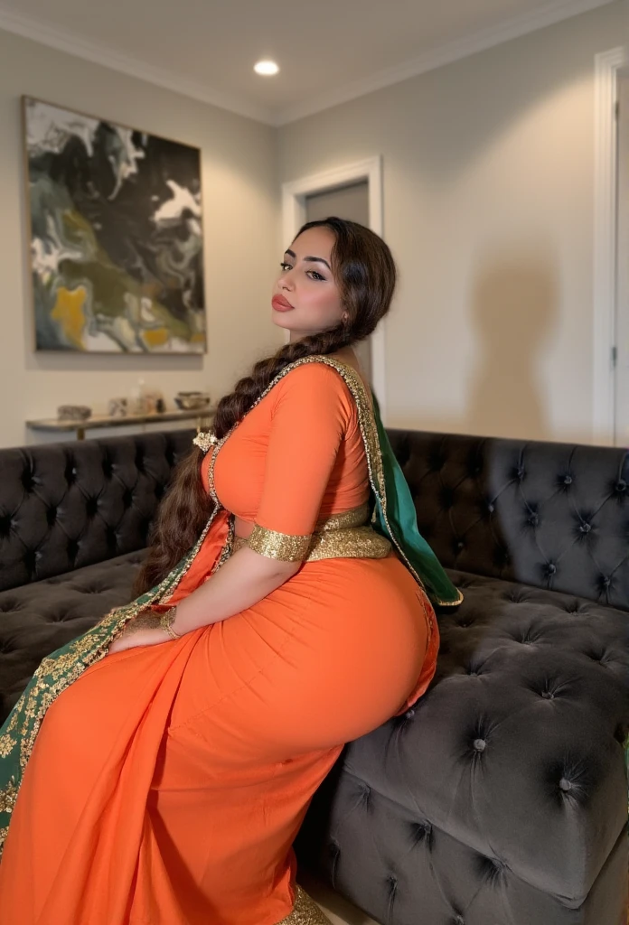 A beautifully curvy woman of South Asian descent sits gracefully on a plush, dark grey tufted couch, adorned in a striking orange saree with intricate embroidery and patterns that highlight her figure. Her saree drapes elegantly around her full, rounded hips and ample curves, emphasizing a voluptuous yet refined silhouette. The blouse fits snugly, subtly hinting at her hourglass shape while leaving her shoulders modestly covered by a rich green dupatta decorated with delicate motifs. Her skin has a warm, light-brown tone that complements the deep, earthy colors of her attire, giving her an aura of traditional elegance.

Her long, thick braid cascades down her back, adorned with a subtle ornament at the end, adding to her graceful charm. She sits with a gentle, soft expression, glancing over her shoulder with a hint of a smile, creating a serene yet subtly inviting look. Behind her, a modern abstract painting and soft, ambient lighting add a layer of intimacy to the scene, blending contemporary and traditional aesthetics in a beautifully balanced way. The setting feels cozy and welcoming, making her presence warm and inviting within the room's classic ambiance.

