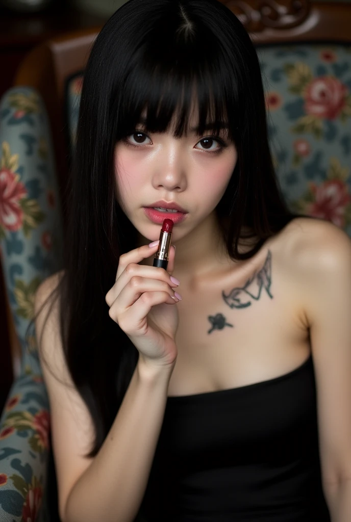 The image is a portrait of a young woman with long black hair and bangs. She is wearing a black strapless dress and has a tattoo on her chest. The woman is holding a lipstick in her right hand and is applying it to her lips. She has a serious expression on her face and is looking directly at the camera. The background appears to be a room with a floral patterned chair. aesthetic posing