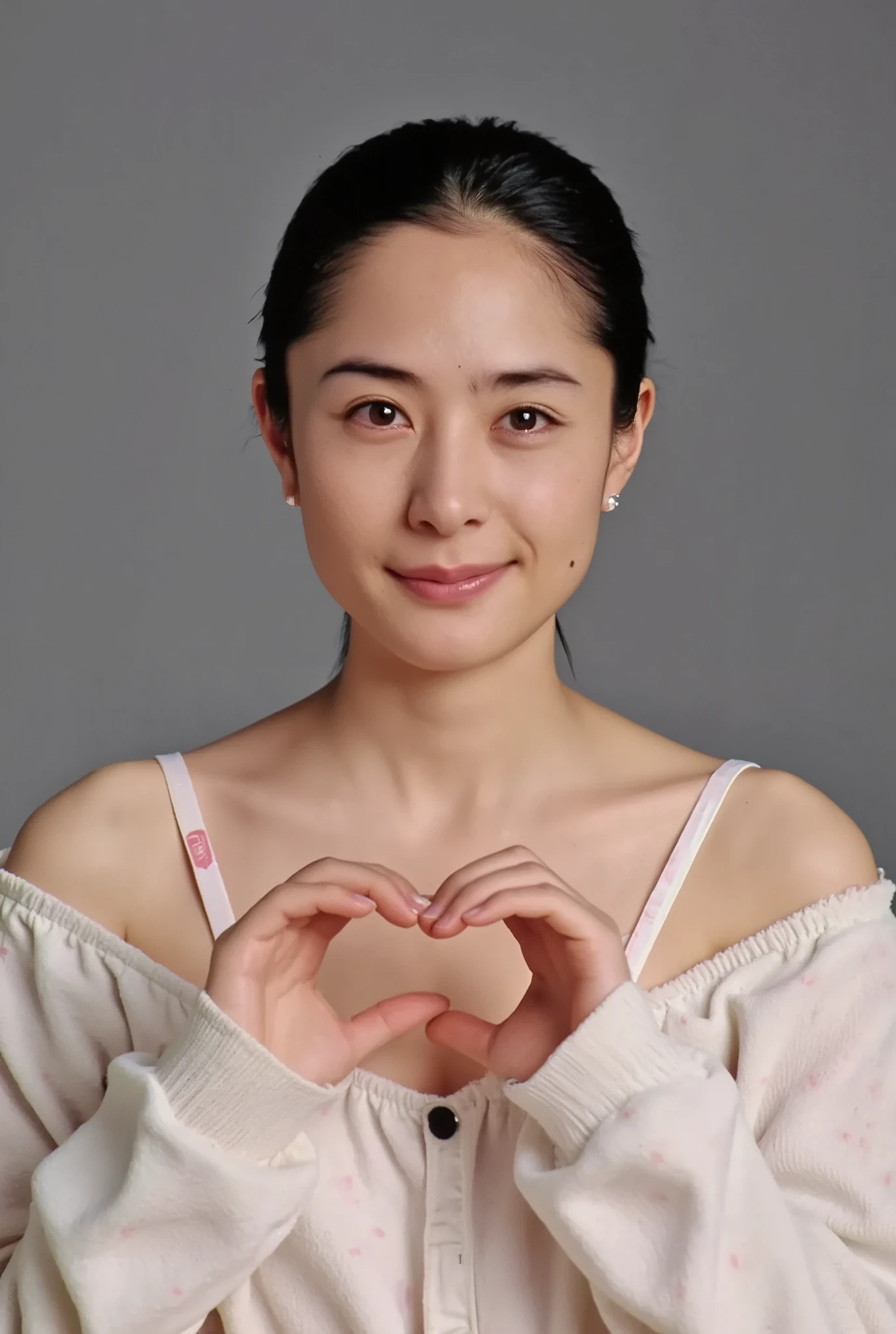 A smiling woman poses alone wearing off-the-shoulder pajamas in pastel colors that convey warmth simply by touching them softly and softly, making a firm, large heart shape with both hands, and holding them in front of her chest, View above collarbone、 background is monotone、
