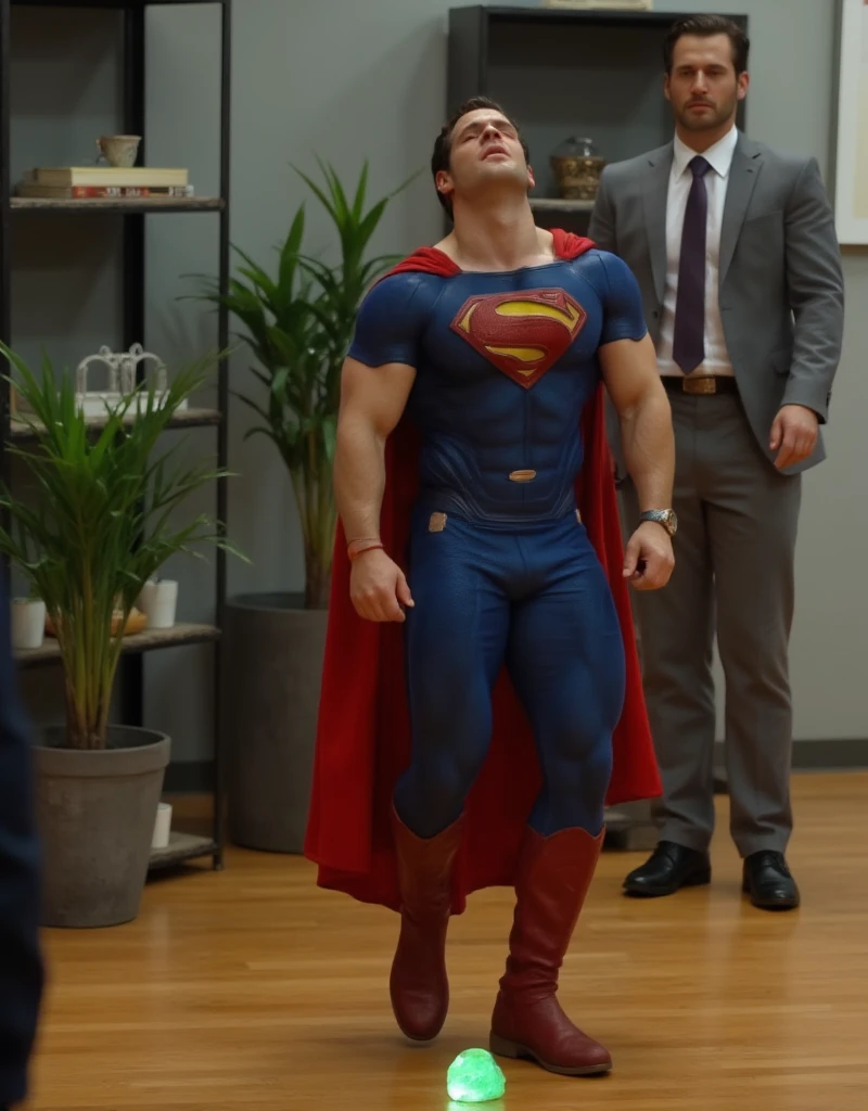 a muscular attractive h3nr4, Henry Cavil with Short stubble in a blue and red superhero costume with a prominent yellow 'S' emblem on the chest, crawling on a wooden floor, orgasm face, eyes closed, mouth open, head down, small glowing green rock on the floor. In the background, there is a blurred view of a modern office setting with shelves and a potted plant. Another person, only partially visible, stands nearby, wearing a gray suit and black shoes