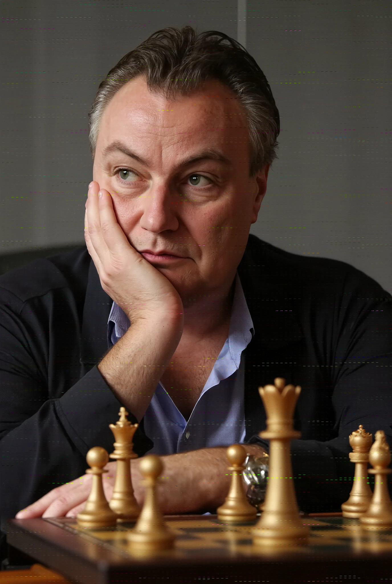 A high-resolution photograph of John Part, sitting behind a chess board, chin resting on his hands, looking down contemplatively,