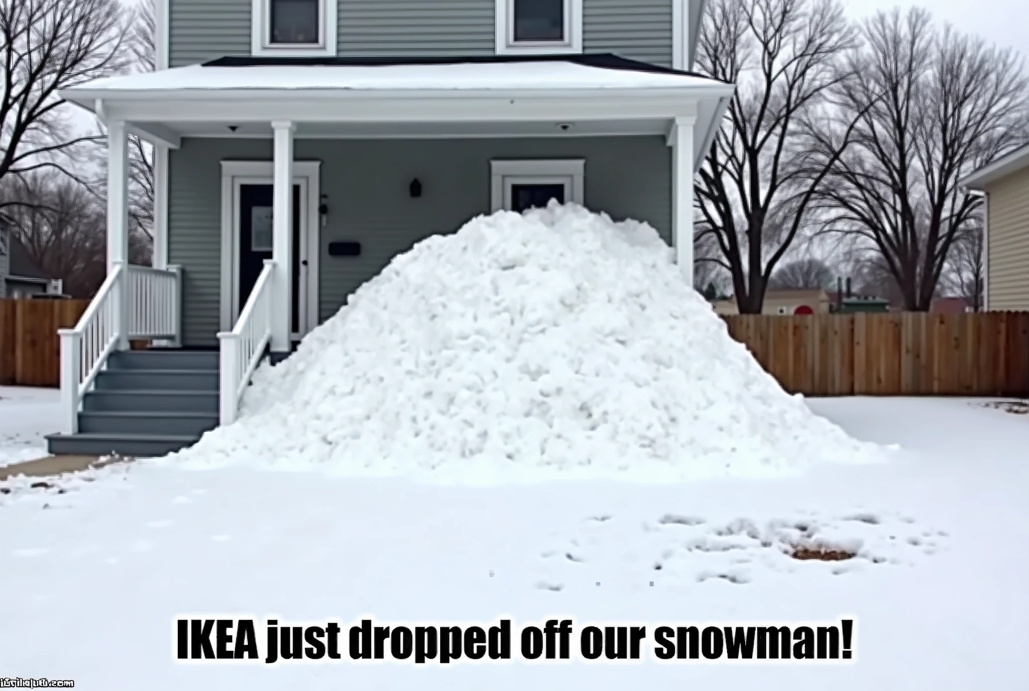 This is a photograph capturing a humorous scene in a snowy residential setting. The image features a large, irregular mound of snow, almost as tall as a small house, which has been dumped just off the front steps of a home. The snow pile is white and fluffy, with a texture that suggests it has been freshly shoveled or dropped from a snowplow. The house is a two-story structure with a gray exterior, featuring white trim and a gray front porch with a set of stairs leading up to the front door. The porch is supported by white pillars and has a small roof overhang. In the foreground, there are several bare patches of ground where the snow has been cleared away, revealing the brown soil underneath. The background shows a line of leafless trees, indicating it is winter, and the sky is overcast, adding to the cold and wintry atmosphere. Text at the bottom of the image reads "IKEA just dropped off our snowman!" in bold, black, sans-serif font, emphasizing the humor of the situation. The overall composition highlights the absurdity of the snow pile, juxtaposed with the everyday setting of a suburban home.