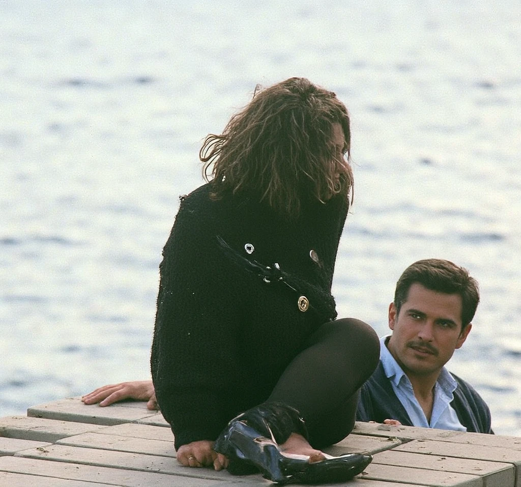 The man in his 45s with black very short hair and smooth face with moustache. The woman in her 45s with brown wavy long hair. The man is wearing an unbuttoned white shirt and black trousers. The woman is wearing a black low-cut evening dress. Seaside, beach, sandy beach. Night. Small ring of fire. The woman is lying on the beach. The man lay on top of the woman. The man penetrating the woman. Sex. Romantic. Passionate. UHD. HDR. Cinematic.