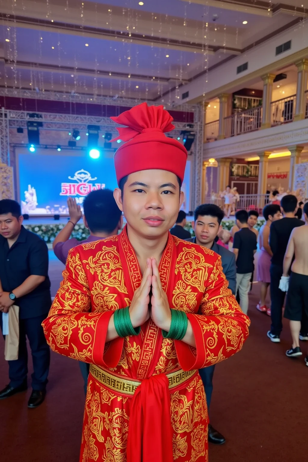 A 30-year-old Thai man stands gracefully in the middle of a luxurious New Year’s celebration, dressed in a traditional Thai outfit styled after the legendary character Gomin. His attire features a vibrant red silk ensemble with intricate patterns, complemented by a matching red sash tied elegantly around his waist. A red silk scarf is tied around his head in a traditional style, adding a bold and iconic touch. He wears jade green bracelets on both wrists, symbolizing strength and cultural heritage. His hands are clasped together in a respectful wai gesture, exuding confidence and pride in his roots. The backdrop is a grand New Year’s event, adorned with shimmering lights, ornate decorations, and a stunningly lit stage. Elegantly dressed guests fill the venue, adding to the festive and sophisticated atmosphere. His composed expression and dignified stance harmonize beautifully with the vibrant and joyous New Year celebration.

