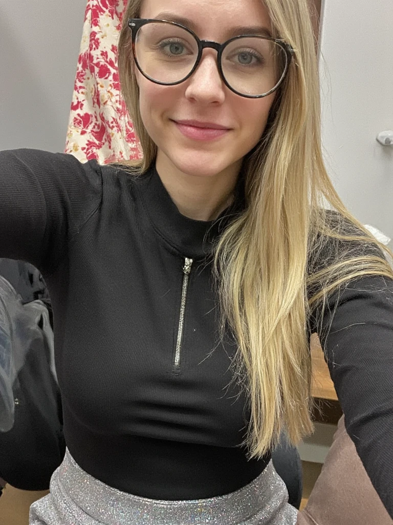 A selfie of a 35-year-old white woman with shoulder-length light blonde hair.  She appears friendly and approachable, with a neutral facial expression.  The woman is wearing a black ribbed, long-sleeved, quarter-zip top.  The top is fitted and has a zippered front closure.  She is wearing a gray, shimmering, skirt-like garment.  The woman is wearing black-framed eyeglasses.  Her skin tone is light. Her pose is casual and slightly angled toward the camera. The lighting in the image is soft and ambient; the color palette is muted and natural, emphasizing the neutral tones of the clothing.  The background consists of a muted gray wall or surface, with a slightly out-of-focus element (possibly a decorative item) with red and white patterns visible partially in the background. The perspective of the image is close, from a slightly above-the-shoulder angle. The texture of the black top is rippled, visible to the ribbed pattern. The texture of the gray skirt is reflective and glittery, showing a slightly sparkly effect,  The overall mood is calm and friendly; the image has a relaxed, informal, and approachable aesthetic. The image has a natural, casual, and slightly intimate feel. Slightly diffused indoor light.  Contemporary style.