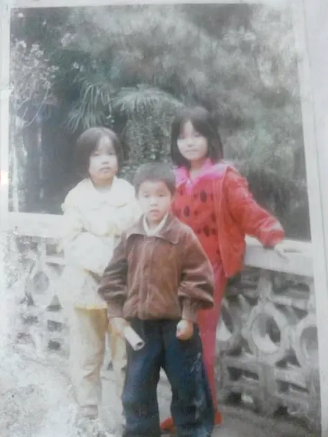 Three small children,The taller boy wears a sweatshirt with a zipper,Another girl wore a white dress,The youngest boy rides a small tricycle,At the front of the car is the head of a sika deer,Stand on the wooden floor,In the background is a lake with houses and trees