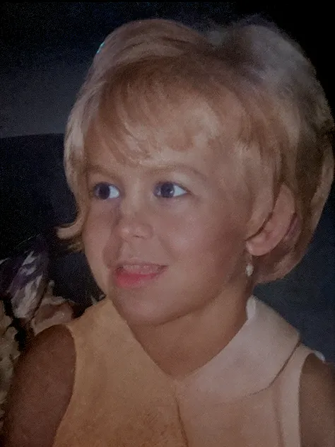 Small with pale, silky, short dark blonde hair with small curls and light blue eyes, taking a photo smiling showing the distracted tongue and the quality of old cameras