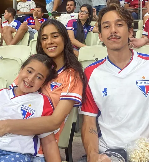 a white woman, with long brown hair, from back with blue jeans and the yellow Colombia shirt, with the number 30 and the name IOMI. On the left, a tall, dark-skinned nine-year-old Latino boy., with short black hair, from back with blue jeans and the yellow Colombia shirt, with the number 04 and the name Sebbas. On the right a 6 year old white boy, with short brown hair, from back with blue jeans and the yellow Colombia shirt, with the number 23 and the name Maximo. The three of them sitting with their backs to each other in a stand in a stadium at night.