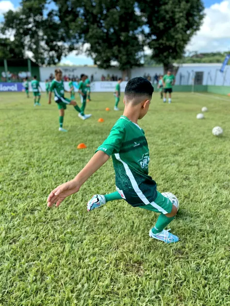 A young Hispanic boy with short black hair enthusiastically playing football. He's wearing a green and white jersey, black shorts, and football boots. His eyes are solely focused on the approaching ball, as he is about to kick it with all his might. His expressive face exhibits determination and passion for the game. The sunlit field in the background is vibrant with green grass, marking lines clearly visible. Players of multiple descents and genders are seen in the distance, preparing to engage in the play once the ball is in motion.