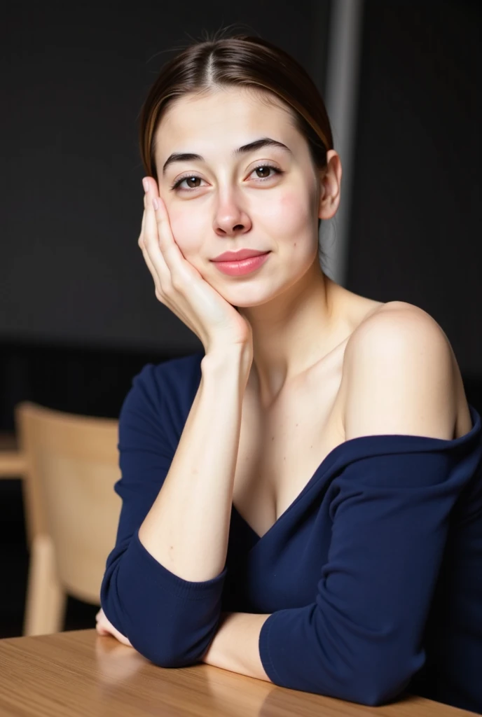 A young woman with blue eyes and dark brown hair in a loose updo, wearing an off-the-shoulder dark blue top, sitting at a table, leaning forward, with one hand touching her chin, soft contemplative expression, dark blurred background
