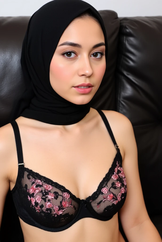 Close-up of a woman wearing a black hijab and a black bra with pink floral embroidery.
The woman is positioned in a slightly angled, three-quarter view, and her face is clearly visible, showing makeup including blush and lipstick. Her expression is neutral.
Her hijab covers her head and shoulders.
The bra is a push-up style with a black lace or mesh base and pink floral appliqués.
The backdrop is a dark, leather-upholstered seat or couch.
Her skin tone appears light to medium.
The image's focus is on the woman's upper body.