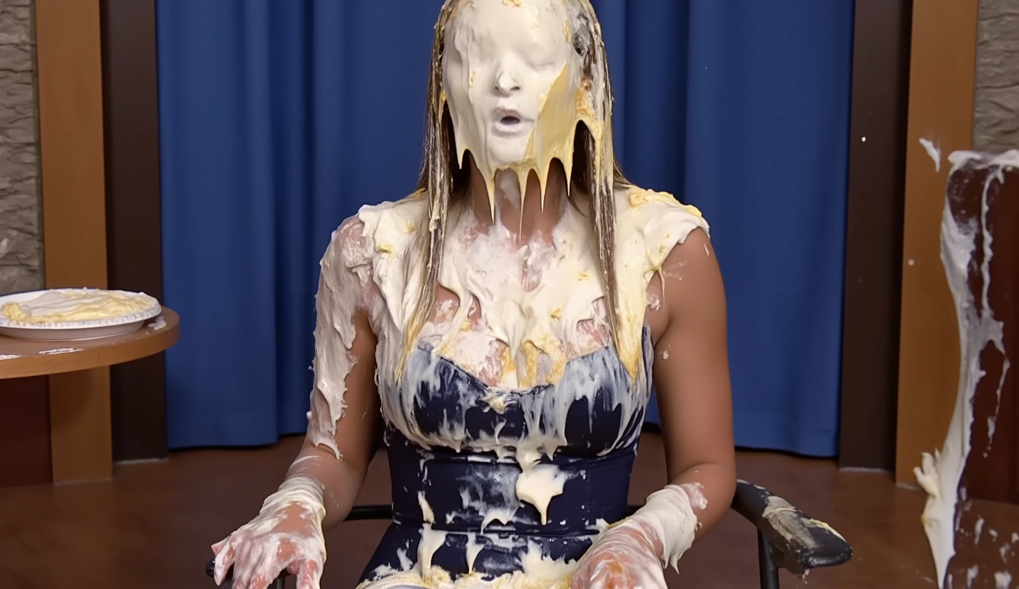 A beautiful young 18-year-old woman with very long, straight blonde hair wearing a dark blue dress sits in a folding chair on the set of a gameshow. She is completely covered from head to toe with very thick white pie cream, including her face, of which her eyes, nose and mouth are completely obscured. Several pie plates are stuck to her. The pies are 100% very thick white cream and have no crusts. They are on white paper plates.