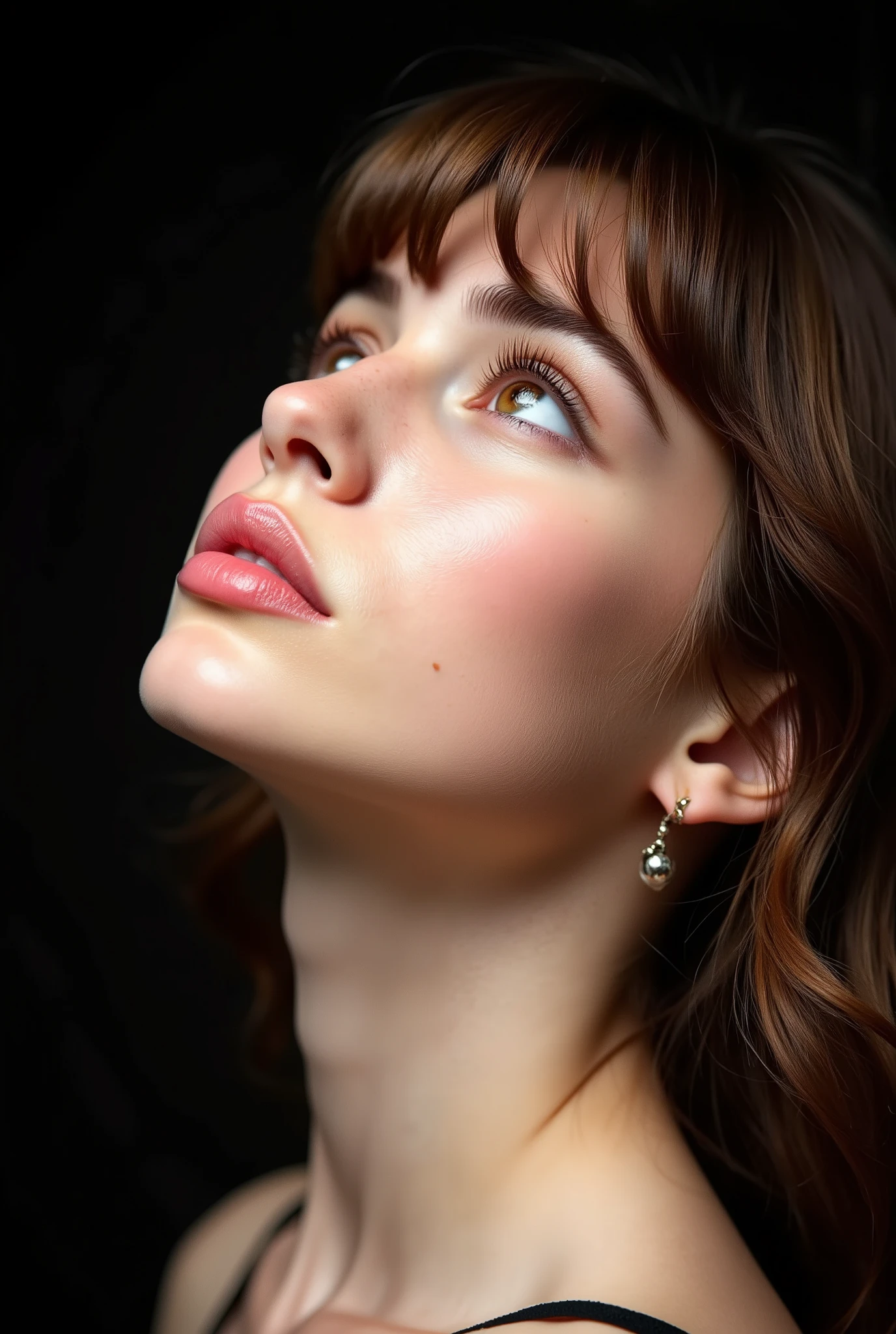 A close-up profile of a woman with fair skin, light pink lipstick, brown eyes, and brown hair in loose waves with strands framing her face, long eyelashes, and defined eyebrows, gazing slightly upward with a neutral expression, against a dark background.